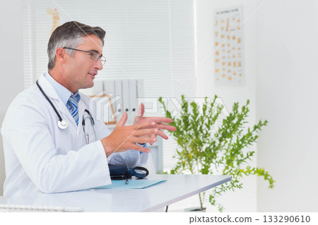 Mature adult male doctor gesturing while explaining diagnosis at desk with stethoscope, copy space Mature adult male doctor gesturing while explaining diagnosis at desk with stethoscope, copy space 133290610