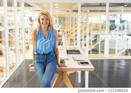 Female office professional sitting on wooden table in open-plan office using monitor and smartphone 133290628