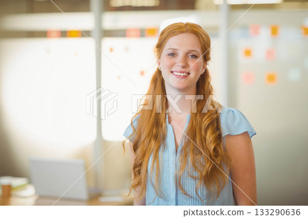 Woman smiling while standing in modern office by frosted glass with sticky notes, laptop and cup 133290636