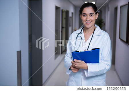 Female doctor standing in hospital hall wearing lab coat with clipboard and stethoscope, copy space Female doctor standing in hospital hall wearing lab coat with clipboard and stethoscope, copy space 133290653