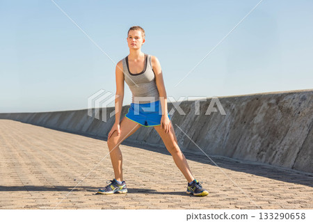 Woman performing lateral stretch on boardwalk by seawall wearing tank top, shorts and running shoes Woman performing lateral stretch on boardwalk by seawall wearing tank top, shorts and running shoes 133290658