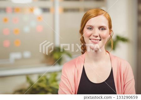 Sitting at desk, woman with red braids wearing coral cardigan near sticky notes and potted plants 133290659