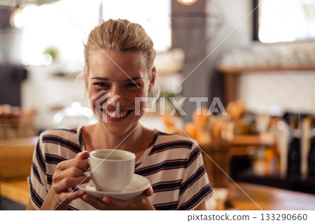Woman smiling and holding ceramic coffee cup at cafe with wicker baskets and jars Woman smiling and holding ceramic coffee cup at cafe with wicker baskets and jars 133290660