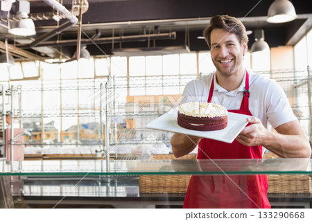 Male baker wearing red apron standing at bakery display counter holding frosted cake, copy space Male baker wearing red apron standing at bakery display counter holding frosted cake, copy space 133290668