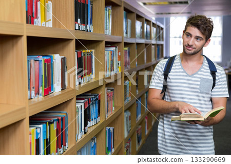 Man standing in library aisle reading open book with backpack under fluorescent lights, copy space Man standing in library aisle reading open book with backpack under fluorescent lights, copy space 133290669
