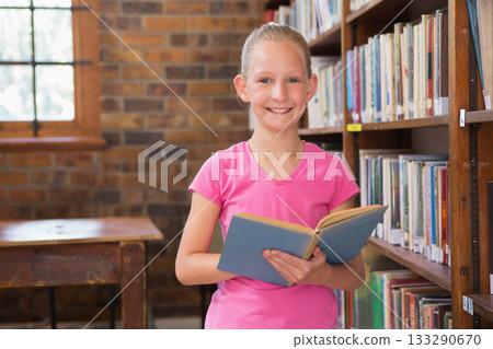 Female child standing in library holding open book and smiling near wooden bookshelf and window Female child standing in library holding open book and smiling near wooden bookshelf and window 133290670