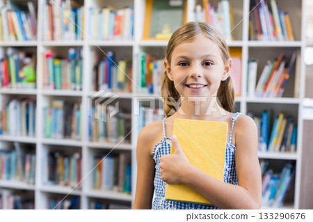 School-age girl in checked dress holding yellow book in library near bookshelves, copy space 133290676