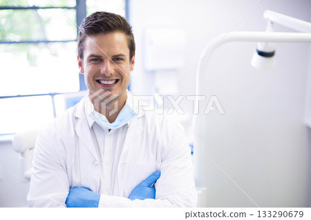 Male dentist standing with crossed arms in dental clinic showing dental light arm and mask 133290679