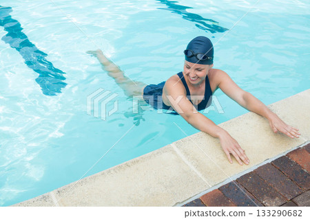 Woman holding edge swimming in pool wearing one-piece swimsuit swim cap goggles over lane markings 133290682