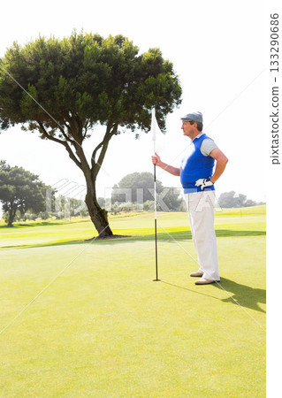 Senior African American man standing on green near tree holding flagstick wearing golf cap, glove 133290686