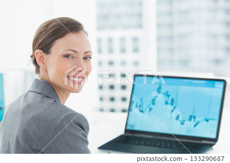 Mid adult woman turning head, smiling at camera at office desk with laptop graph, copy space Mid adult woman turning head, smiling at camera at office desk with laptop graph, copy space 133290687