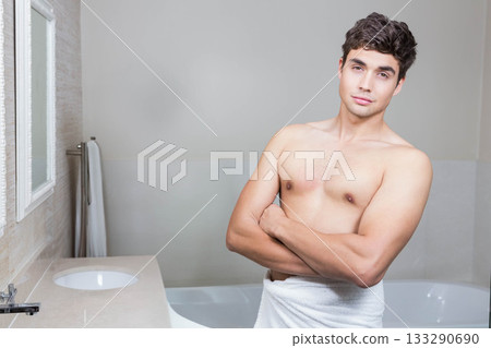 Man in twenties wrapped in white towel standing in bathroom by sink and bathtub, copy space Man in twenties wrapped in white towel standing in bathroom by sink and bathtub, copy space 133290690