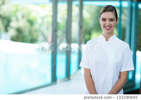 Woman in white tunic standing in bright corridor overlooking pool through glass panels, copy space 133290695