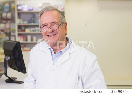 Senior male pharmacist in lab coat standing behind counter at pharmacy using computer monitor Senior male pharmacist in lab coat standing behind counter at pharmacy using computer monitor 133290704