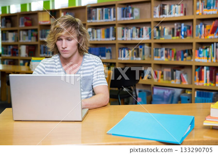 Male student reading on laptop at wooden library table with blue binder and books 133290705