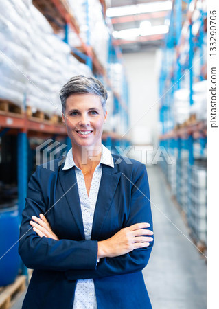 Mature adult woman standing in warehouse aisle wearing navy blazer amid steel racks of white sacks 133290706