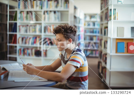 Teenage boy sitting at round table in library reading open textbook with pen and holding smartphone 133290724