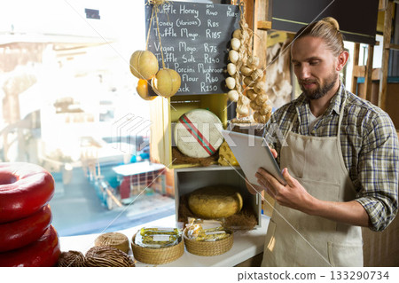 Male shopkeeper holding tablet, showcasing cheese wheels, baskets on shop counter, copy space 133290734