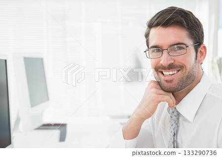 Businessman smiling and resting chin on hand at office desk with monitor, keyboard, copy space Businessman smiling and resting chin on hand at office desk with monitor, keyboard, copy space 133290736