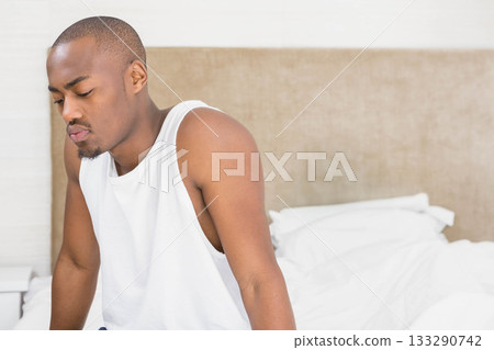 African American man sitting on edge of bed in bedroom examining rumpled white sheets and pillows 133290742
