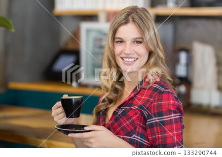 Woman in her twenties holding cup and saucer next to espresso machine on wooden cafe counter Woman in her twenties holding cup and saucer next to espresso machine on wooden cafe counter 133290744