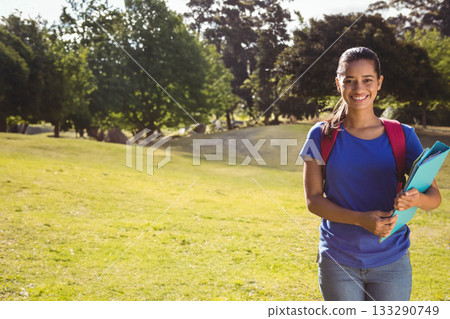 Female student standing on lawn holding notebooks and folders wearing red backpack, copy space 133290749