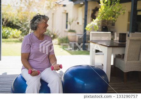 Senior woman balancing on blue exercise ball on wooden patio deck holding pink dumbbells 133290757