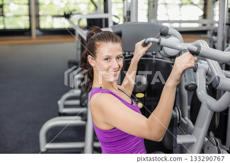 Woman wearing purple tank top and black leggings using chest press machine at fitness center 133290767
