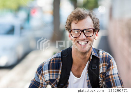 Man standing and smiling on sunlit city sidewalk while wearing plaid shirt, glasses and backpack Man standing and smiling on sunlit city sidewalk while wearing plaid shirt, glasses and backpack 133290771