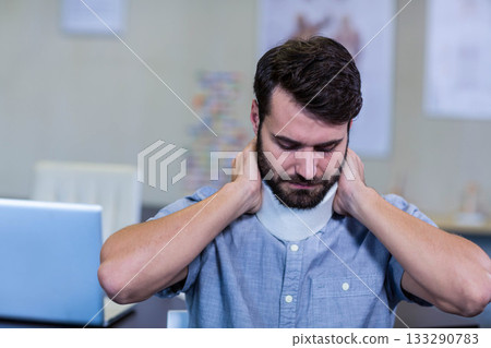 Bearded man in light blue shirt adjusting cervical collar support at desk in office with laptop 133290783
