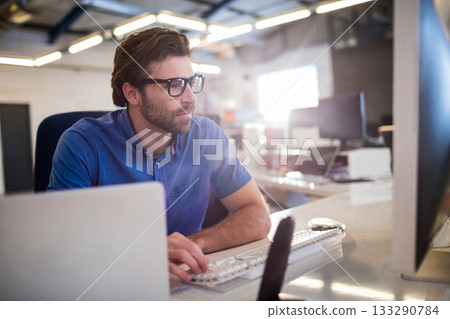 Man in his thirties in blue shirt typing on wireless keyboard at office desk with laptop Man in his thirties in blue shirt typing on wireless keyboard at office desk with laptop 133290784