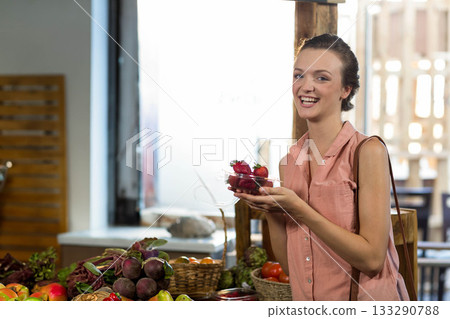 Woman holding plastic container of strawberries and smiling at market stall among wooden crates 133290788