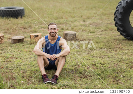 Adult male fitness enthusiast in blue top sitting, smiling on field with tractor tires and stumps Adult male fitness enthusiast in blue top sitting, smiling on field with tractor tires and stumps 133290790