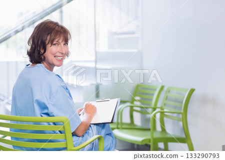 Female nurse sitting on green chair in waiting room writing on clipboard smiling back, copy space Female nurse sitting on green chair in waiting room writing on clipboard smiling back, copy space 133290793