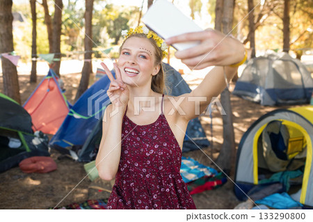 Woman wearing floral headband taking selfie using smartphone at forest camp with tents and bunting 133290800