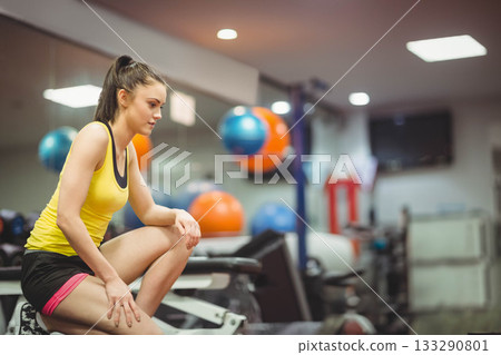 Woman in sportswear sitting on weight bench raising foot in gym with stability balls, copy space Woman in sportswear sitting on weight bench raising foot in gym with stability balls, copy space 133290801