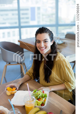 Female office employee smiling while holding small fruit container at cafeteria table with salad Female office employee smiling while holding small fruit container at cafeteria table with salad 133290804