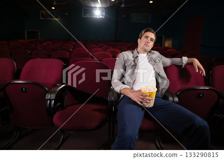 Man slouching in red theater seat under projection beam holding yellow popcorn bucket, copy space 133290815