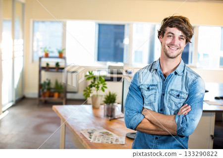 Man standing cross-armed in open office space by wooden desk with laptop and plants, copy space Man standing cross-armed in open office space by wooden desk with laptop and plants, copy space 133290822