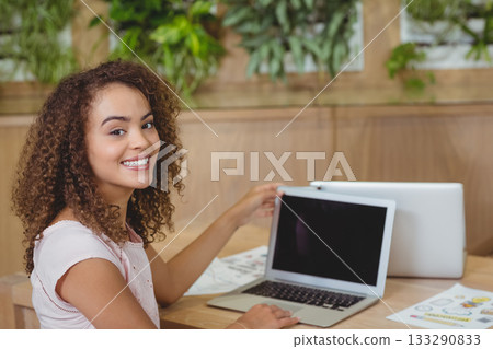 Female professional turning laptop screen toward viewer at wooden desk in coffee shop with papers Female professional turning laptop screen toward viewer at wooden desk in coffee shop with papers 133290833