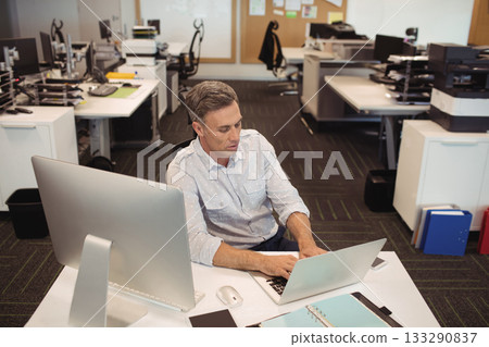Middle-aged man in business casual attire typing on laptop at desk in open-plan office with monitor 133290837