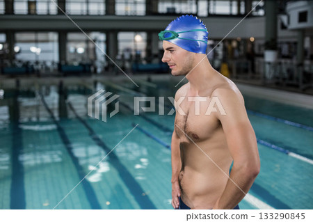 Male swimmer standing on lap-pool deck by lane ropes wearing blue cap, goggles, briefs, copy space 133290845