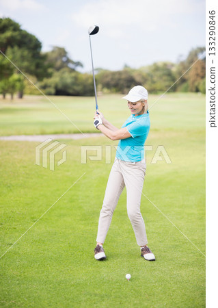Female golfer swinging driver on golf course fairway with cap glove and golf ball on tee 133290846