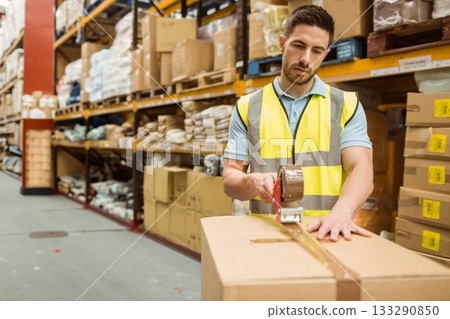 Man wearing safety vest sealing cardboard box with tape dispenser in warehouse aisle, copy space 133290850