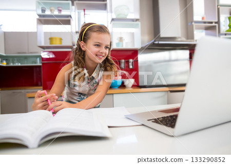 School-age girl studying homework at kitchen counter with laptop, textbook, pen, and mixing bowls 133290852