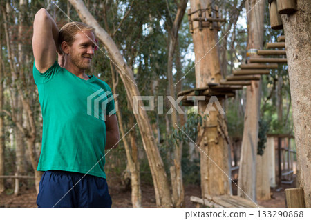 Mid adult man stretching triceps in green shirt at ropes course amid rope bridges, copy space 133290868