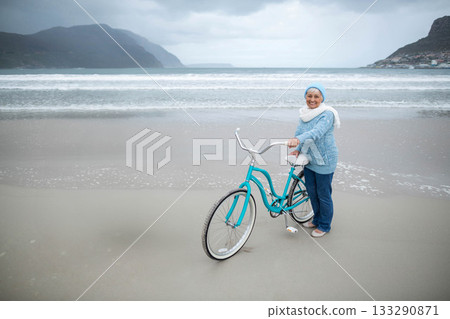Senior female holding turquoise bicycle standing on wet sand at shore under cloudy sky, copy space 133290871
