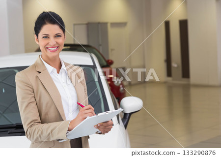 Woman in blazer standing beside white car in showroom holding clipboard with red pen, copy space 133290876