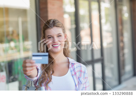 Woman in her twenties smiling while holding credit card forward on sidewalk by storefront window Woman in her twenties smiling while holding credit card forward on sidewalk by storefront window 133290888