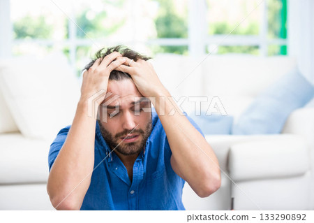 Clutching head with hands, man in blue shirt sitting on floor at home by couch, window 133290892